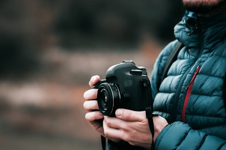Close-up image of a photographer holding a DSLR camera outdoors in a puffer jacket, showcasing focus and creativity.